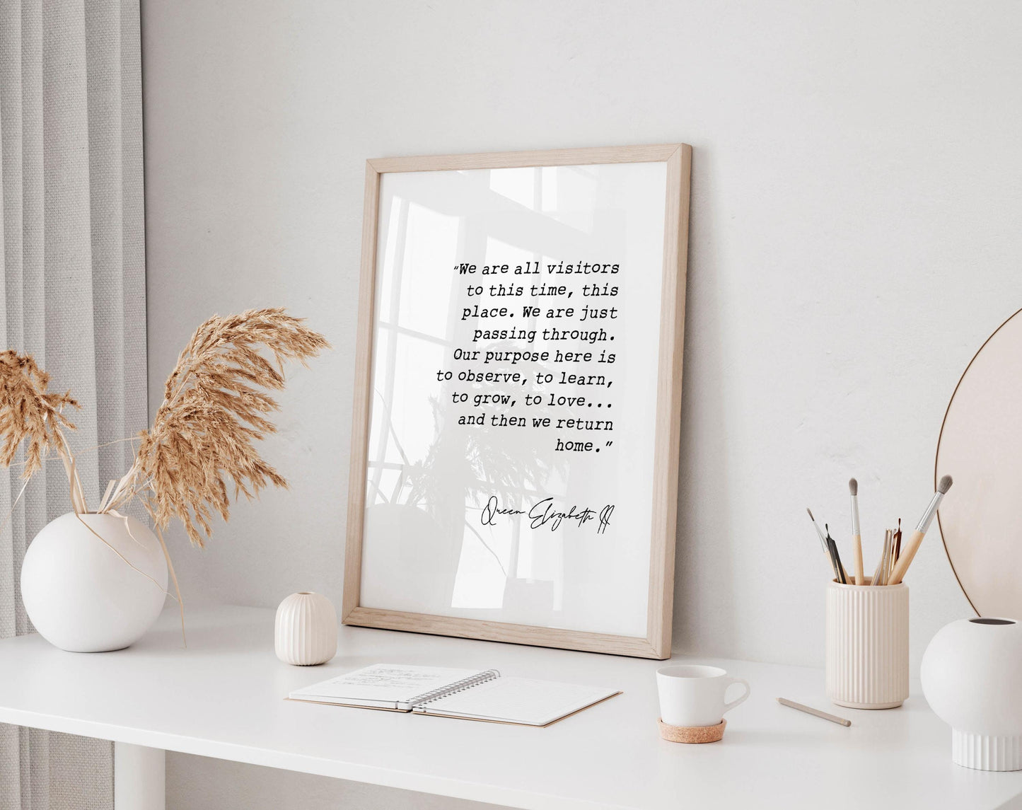a framed quote on a white wall, accompanied by a small notebook, a white mug, and a white vase with dried flowers.