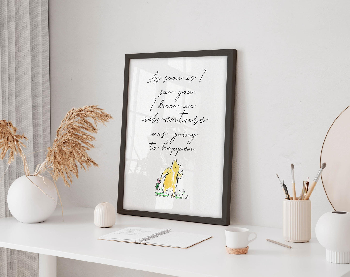 a framed quote on a white desk, surrounded by various objects such as a notebook, a cup, and a vase with dried flowers.