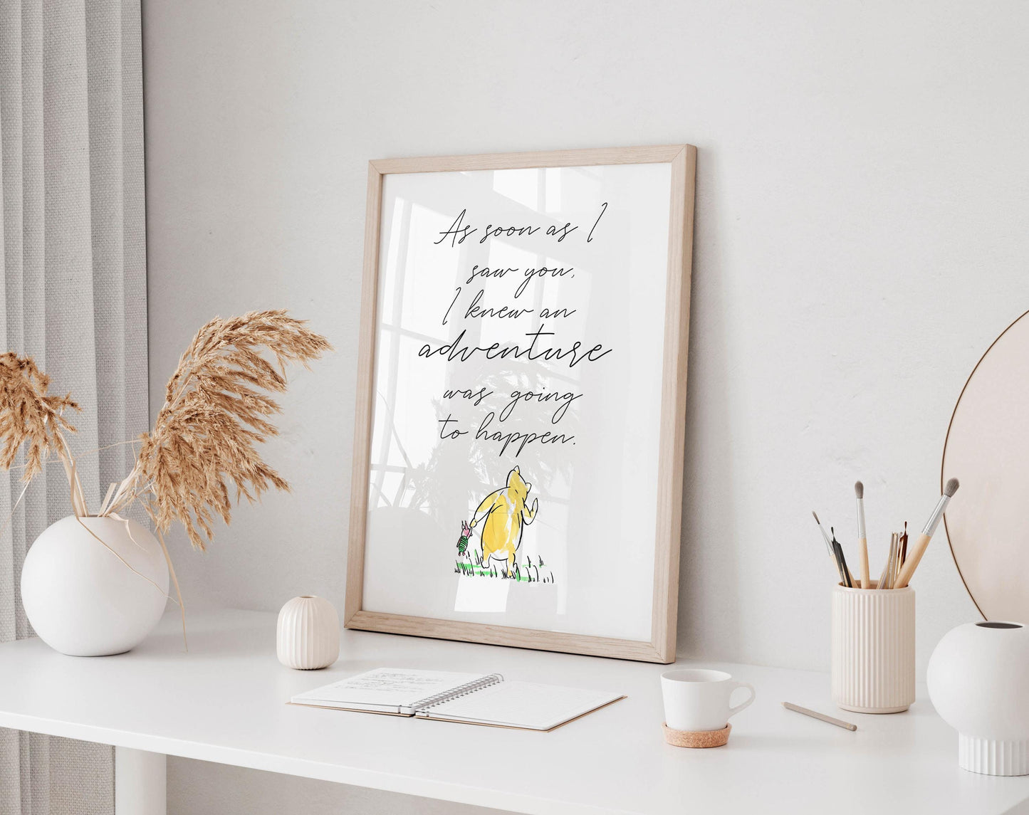 a framed quote on a white desk, surrounded by various objects such as a notebook, a cup, and a vase with dried flowers.