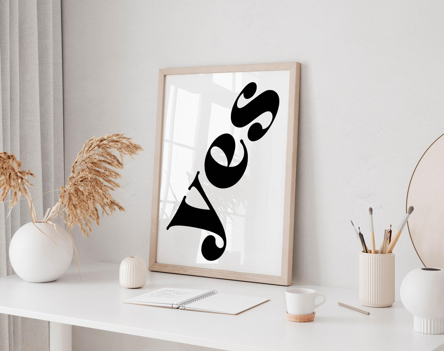 a white desk with a framed poster displaying the word &quot;YES&quot; in bold black letters, a small white vase with dried flowers, a white mug, a notebook, and a small white figurine.