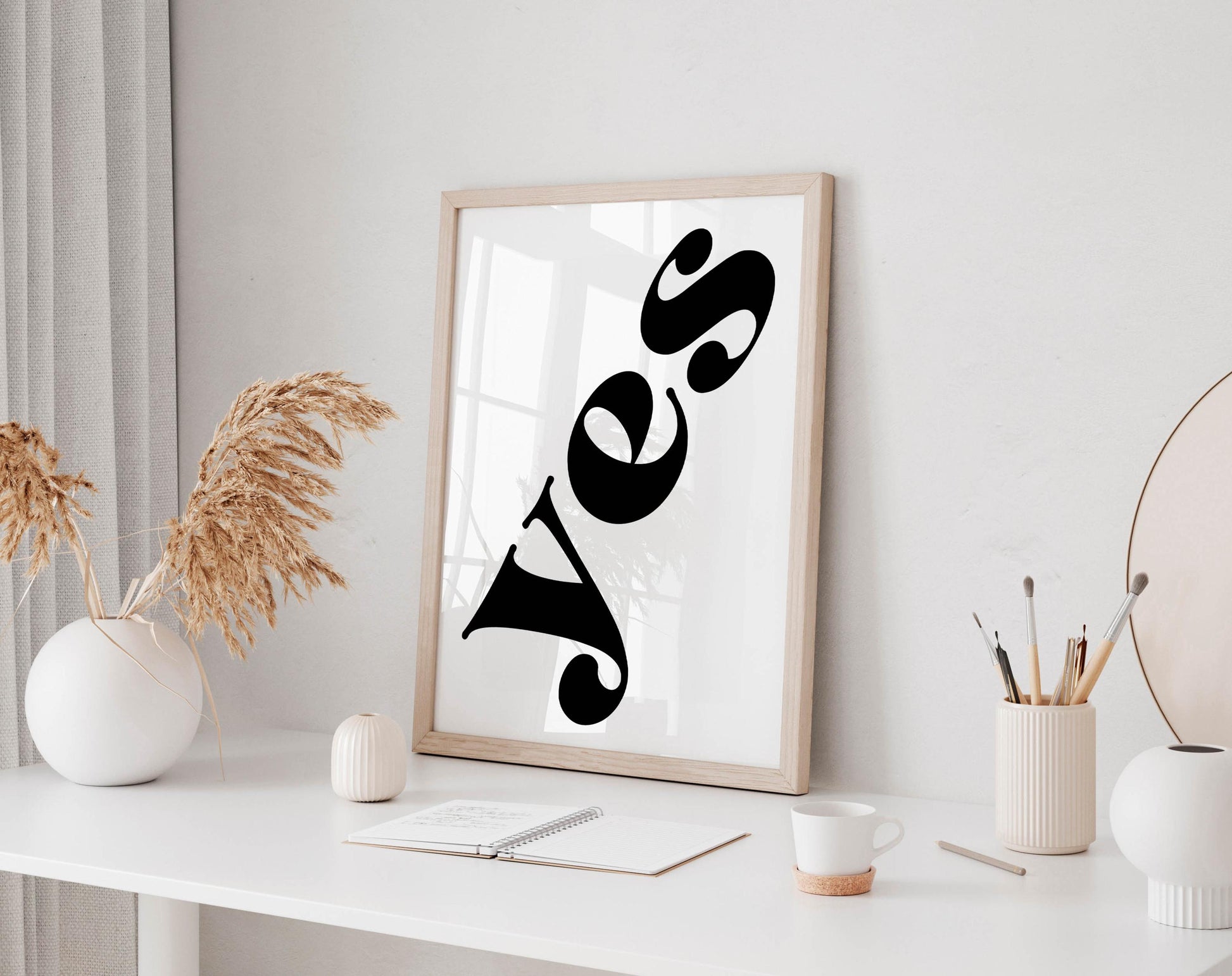 a white desk with a framed poster displaying the word &quot;YES&quot; in bold black letters, a small white vase with dried flowers, a white mug, a notebook, and a small white figurine.