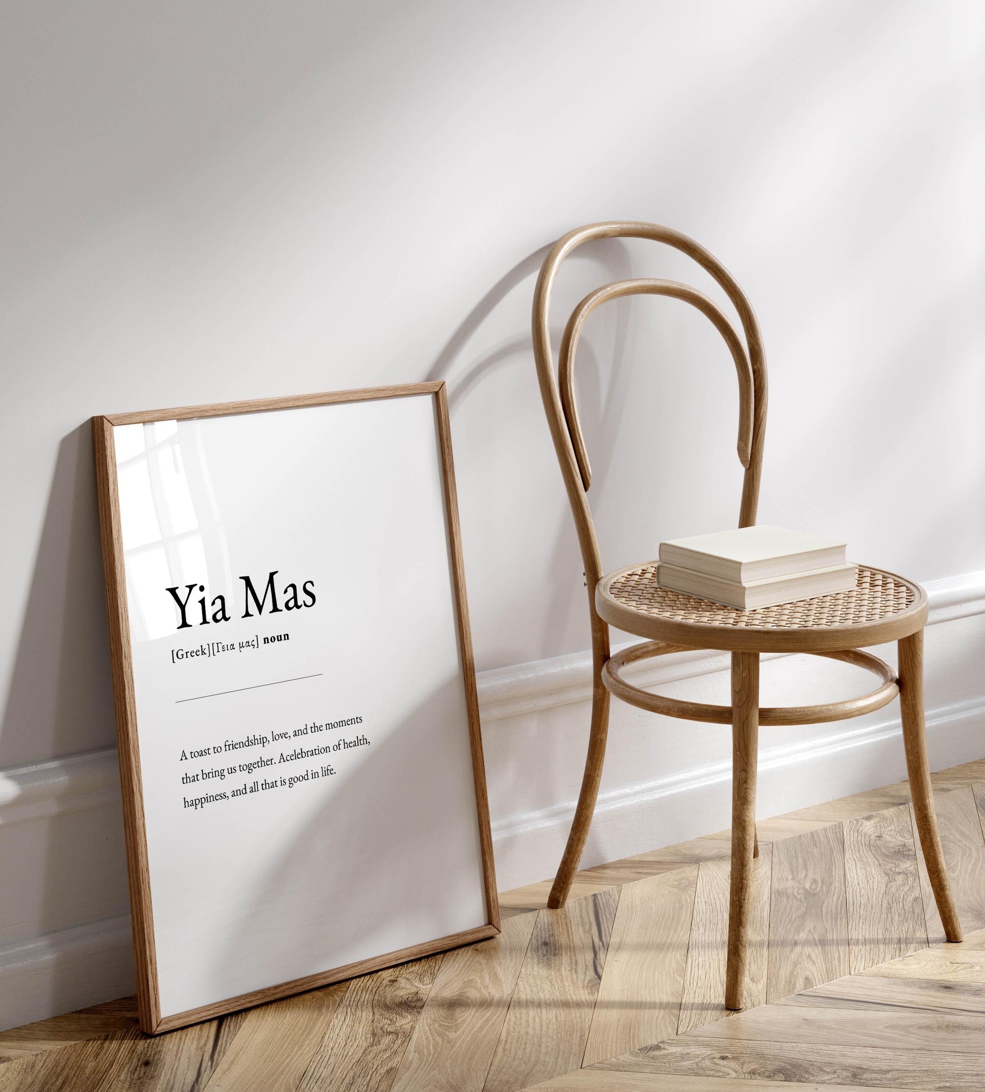 A white framed definition of &#39;Yia Mas&#39; is leaning against a white wall, accompanied by a wooden chair with a stack of books on it.