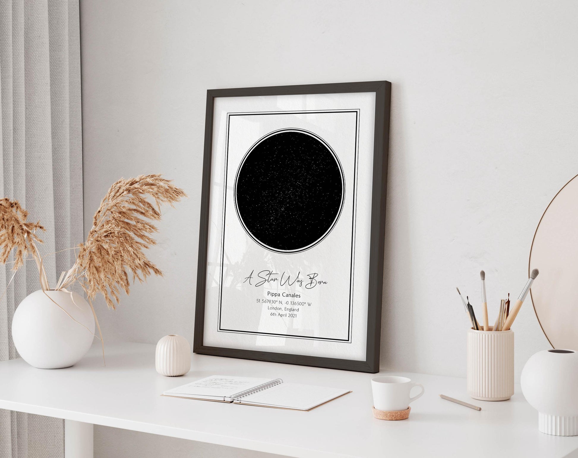 a framed picture of a starry night sky on a white desk, accompanied by a notebook, a cup, and a vase with dried flowers.