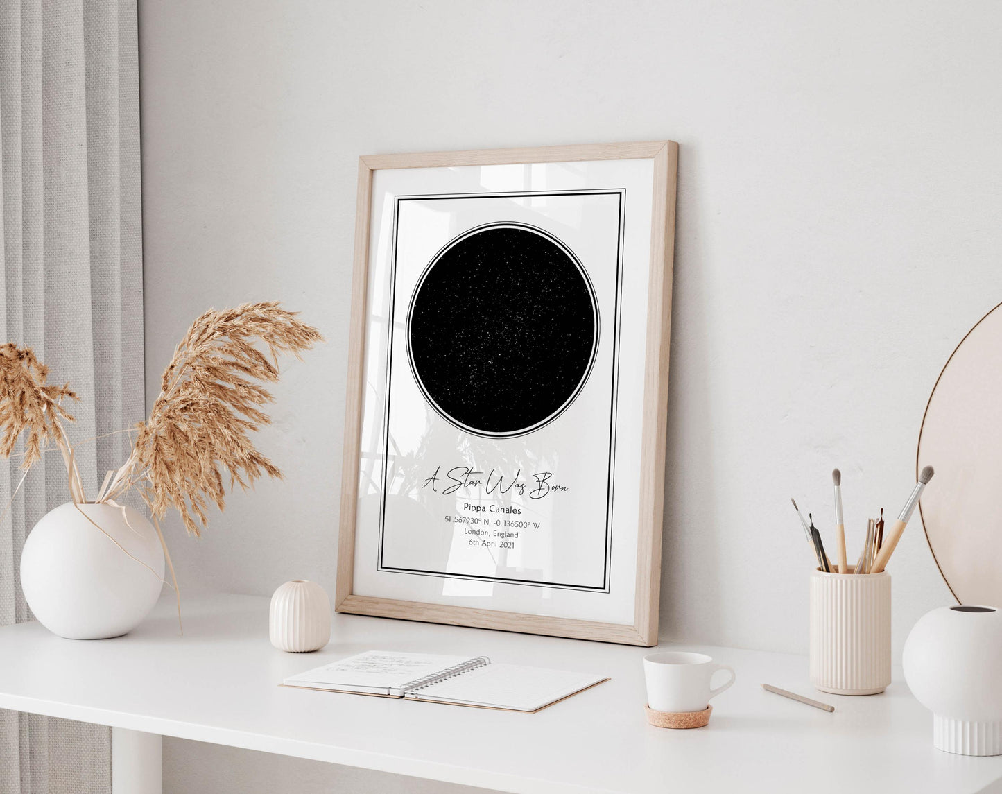 a framed picture of a starry night sky on a white desk, accompanied by a notebook, a cup, and a vase with dried flowers.