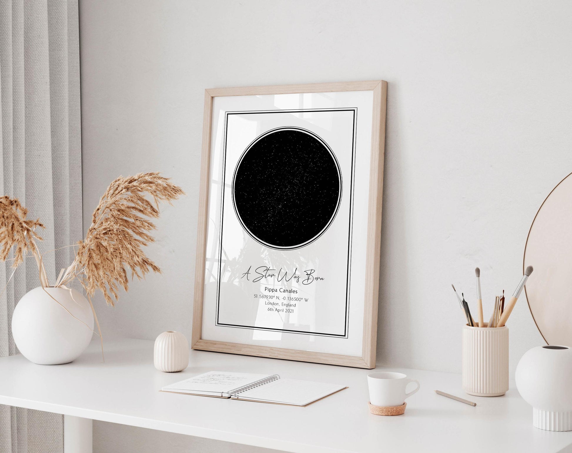 a framed picture of a starry night sky on a white desk, accompanied by a notebook, a cup, and a vase with dried flowers.