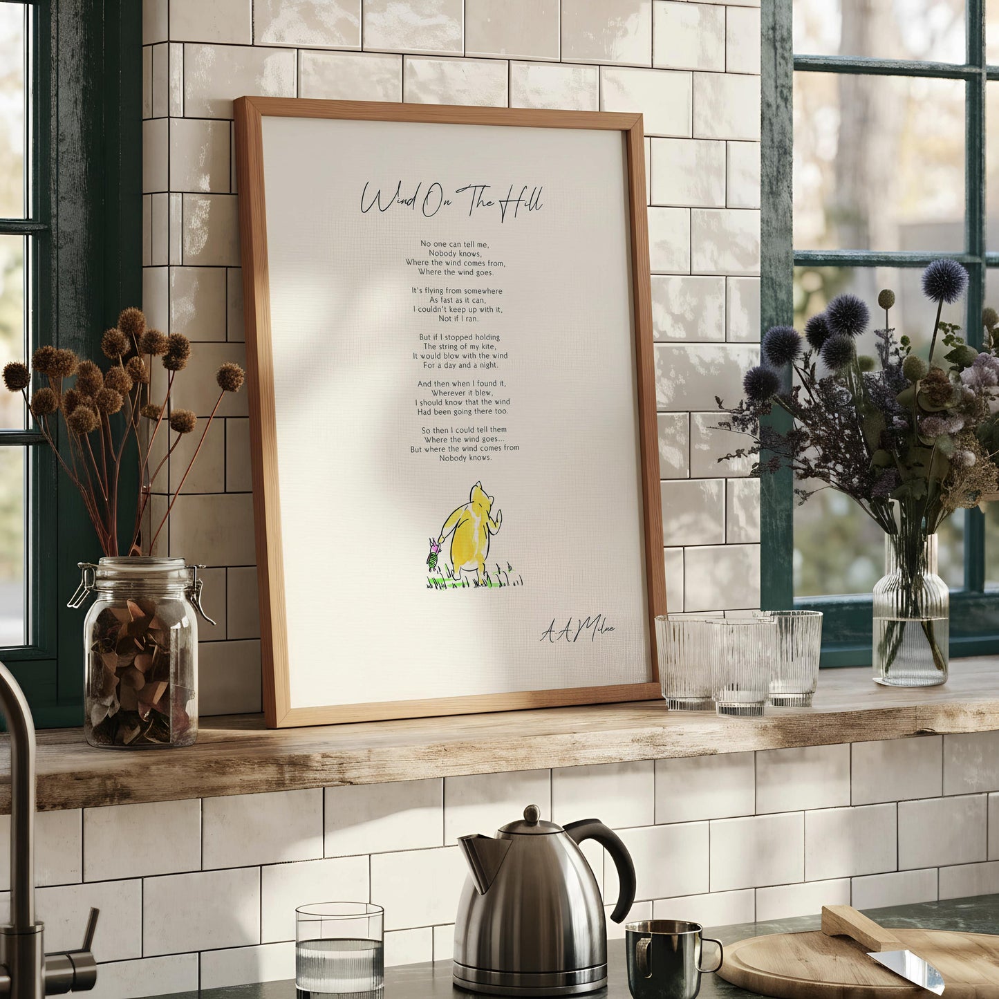 a framed poem titled "Wind on the Hill" by A. A. Milne, which is a children's book by the same author. The poem is set against a white background and is framed in a wooden frame. The text of the poem is displayed in a clear, legible font.