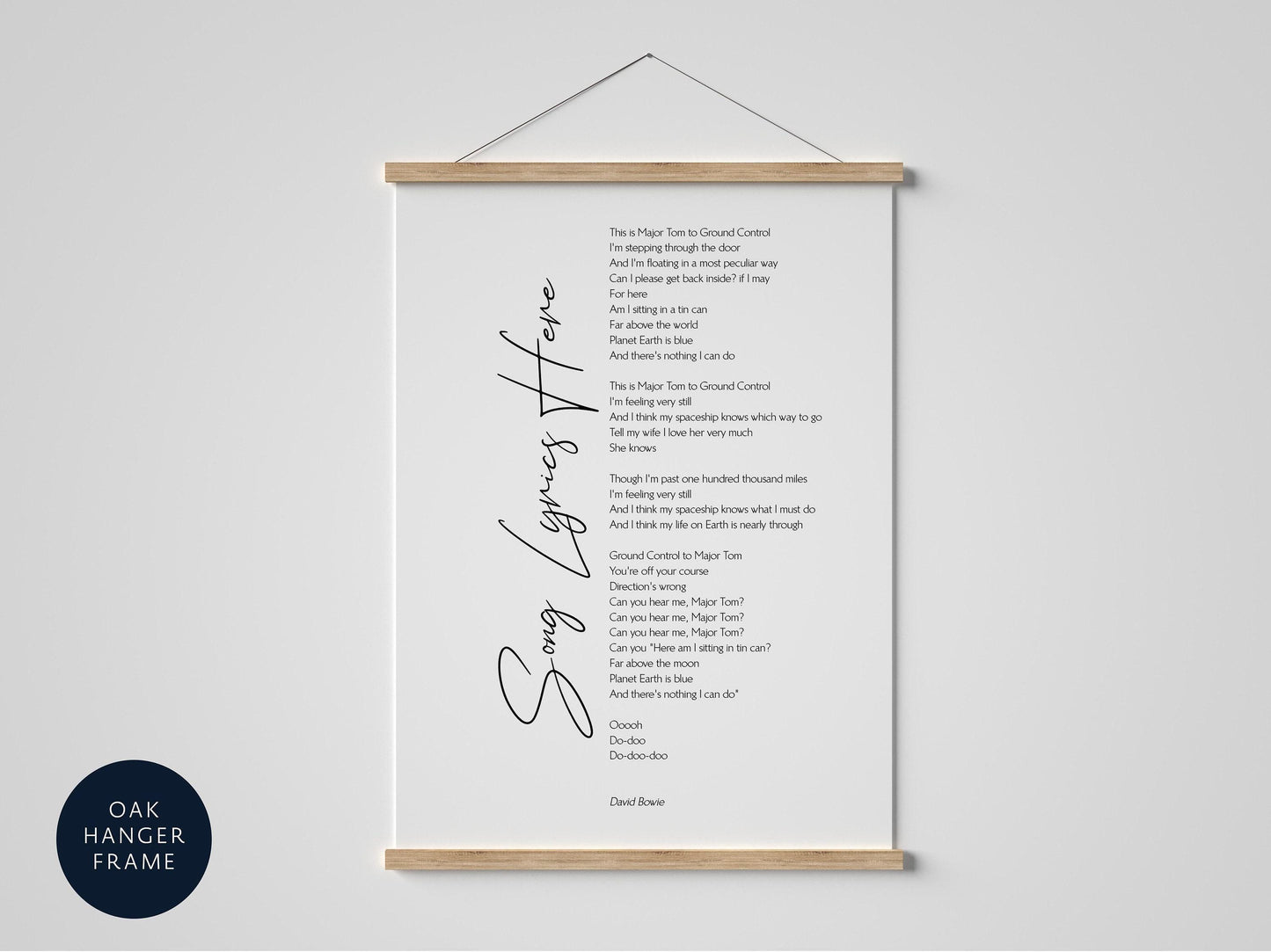 May include: A white poster featuring the lyrics of David Bowie's "Space Oddity" in black text, held by an oak hanger frame. The poster hangs against a white wall. The words "Song Lyrics Here" are written in cursive. The frame is made of wood.