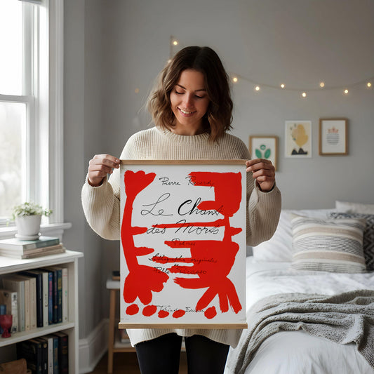 a framed poster with a red and white abstract design, placed on a white desk next to a white vase with dried flowers and a white mug.