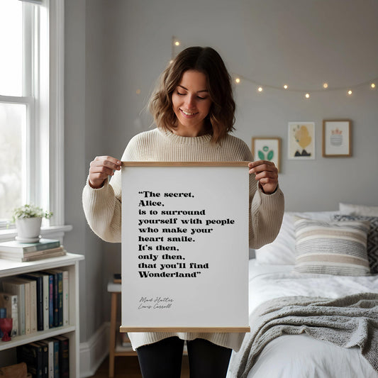 a framed quote by Lewis Carroll, "The secret, Alice, is to surround yourself with people who make your heart smile. It's then, only you'll find Wonderland." The quote is displayed on a white wall, accompanied by a small notebook, a white mug, and a white vase with dried flowers.