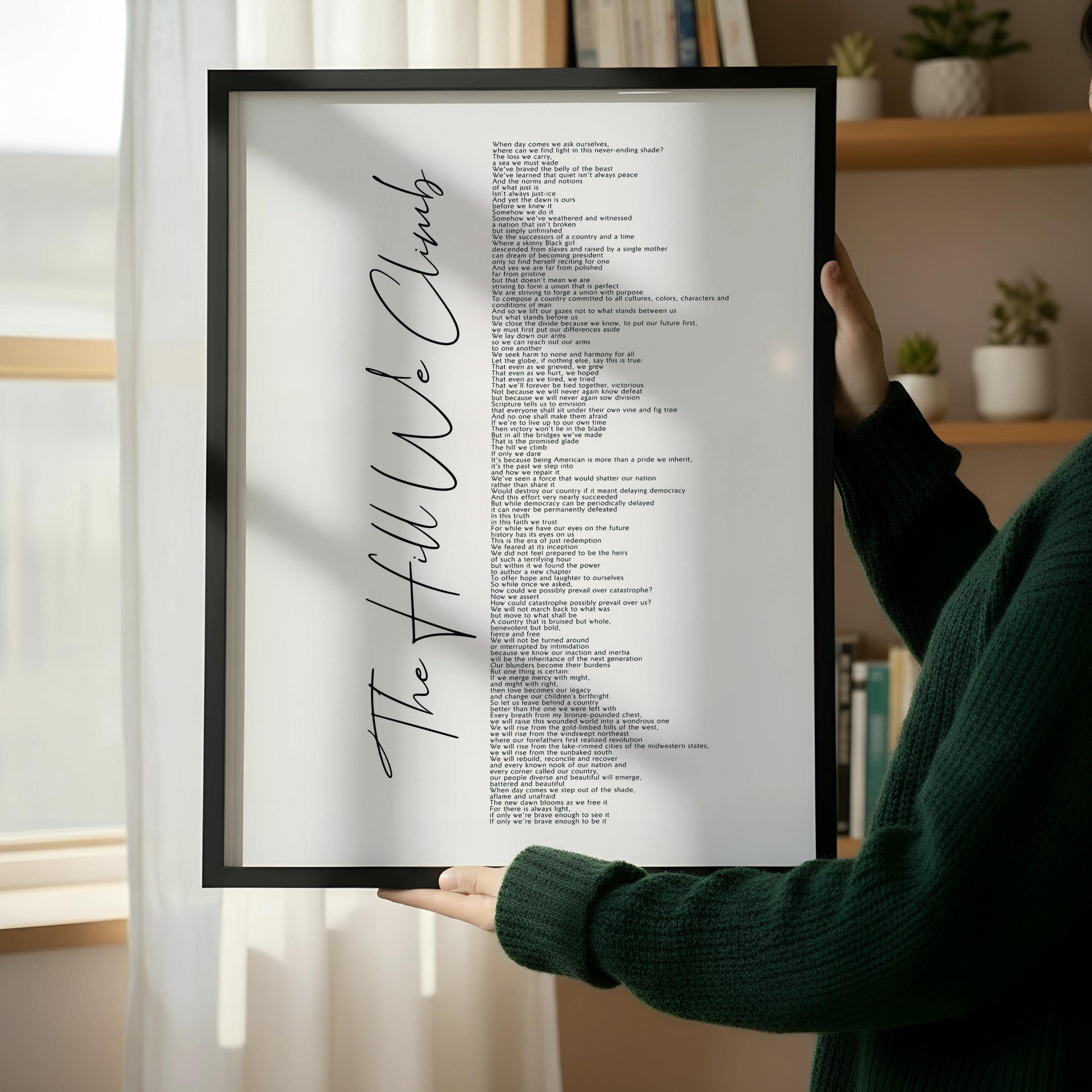 The image is a framed print with a list of words written in black cursive font, which appears to be a song or poem. The words are arranged in a vertical format, with the title "The Hill We Climb" written at the top. The frame has a wooden border, and the background of the image is a light color.