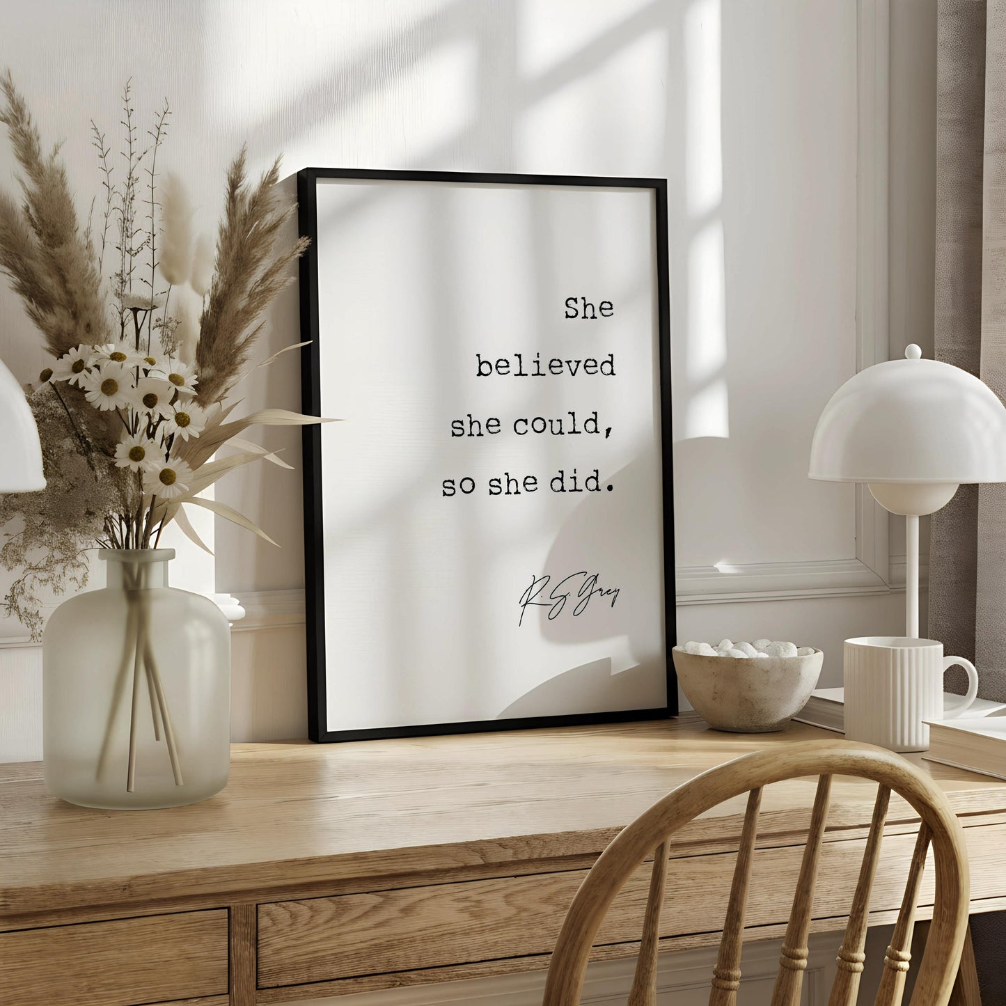 A framed motivational quote is displayed on a white desk, accompanied by a notebook, a cup, and a vase with dried flowers.