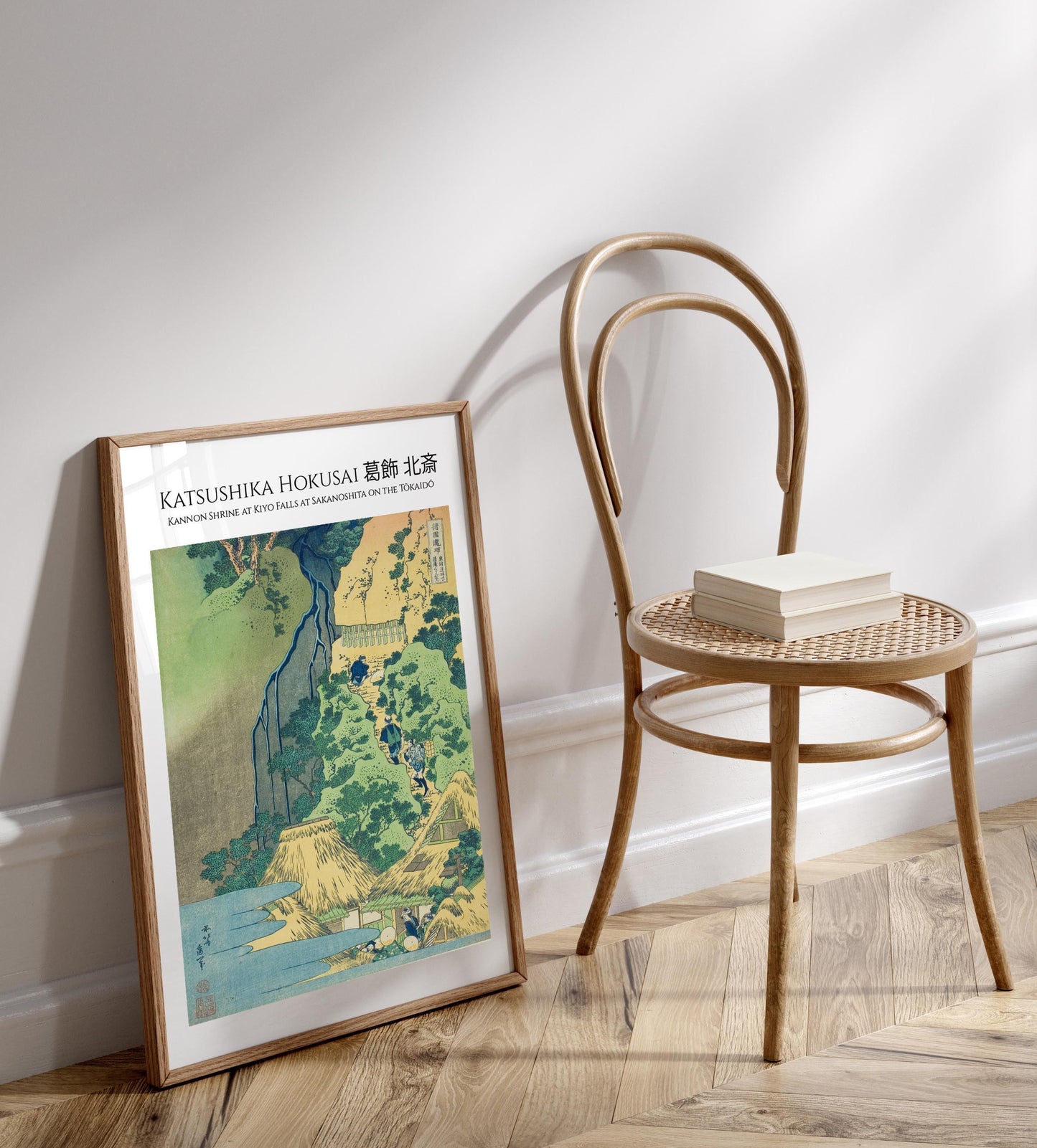a framed print of a landscape painting titled "Katsushika Horosasa" by the artist Kanno Shinshire, which depicts a waterfall and a temple. The painting is hanging on a wall next to a wooden chair, and there is a book placed on the chair.
