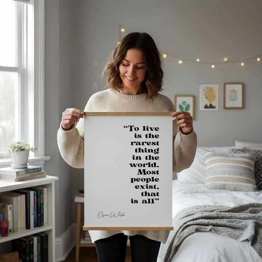 a framed quote by Oscar Wilde, "To live is the rarest thing in the world. Most people exist, that is all", placed on a white desk with a notebook, pen, and a cup of coffee.