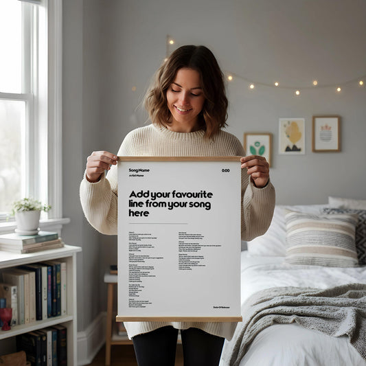 May include: A white poster with a wooden frame, displaying the text "Add your favourite line from your song here" in black. The poster includes song lyrics and artist details. The poster is held by a person.