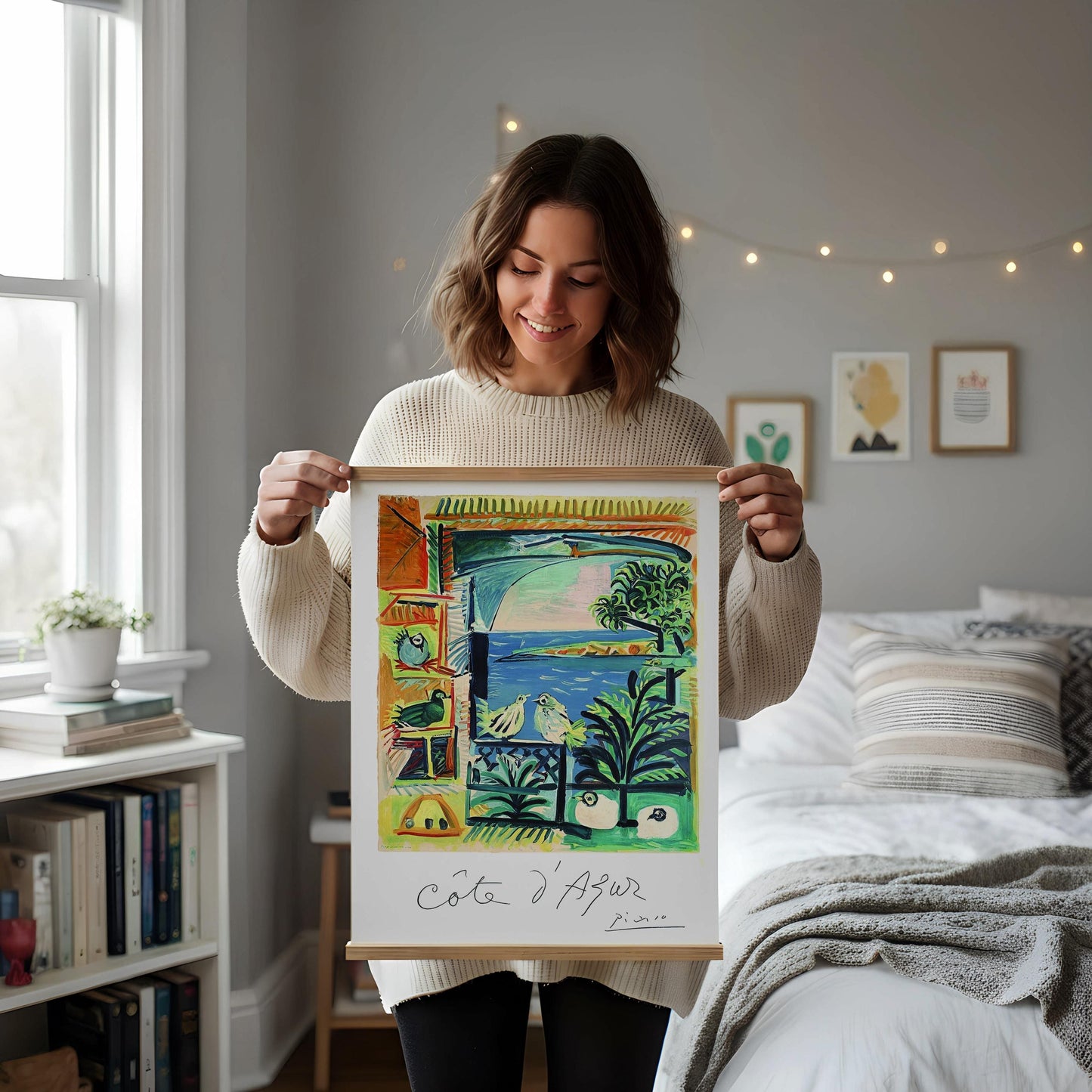 a framed painting of a tropical landscape with palm trees, birds, and a body of water, displayed on a white shelf. The painting is surrounded by various decorative items, including a vase with dried flowers, a small white candle, and a white mug.