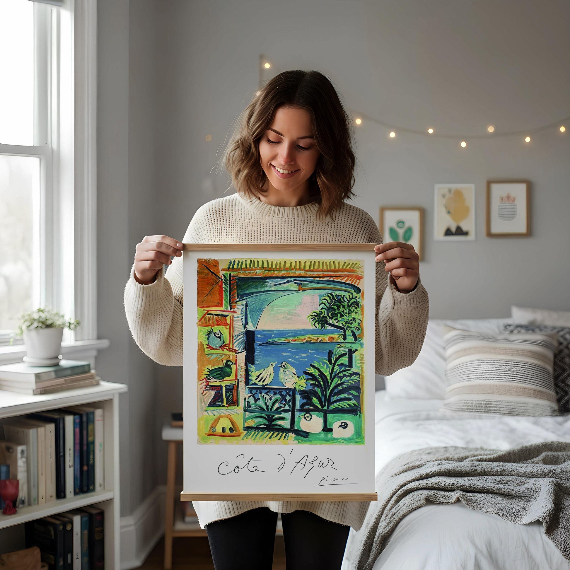 a framed painting of a tropical landscape with palm trees, birds, and a body of water, displayed on a white shelf. The painting is surrounded by various decorative items, including a vase with dried flowers, a small white candle, and a white mug.