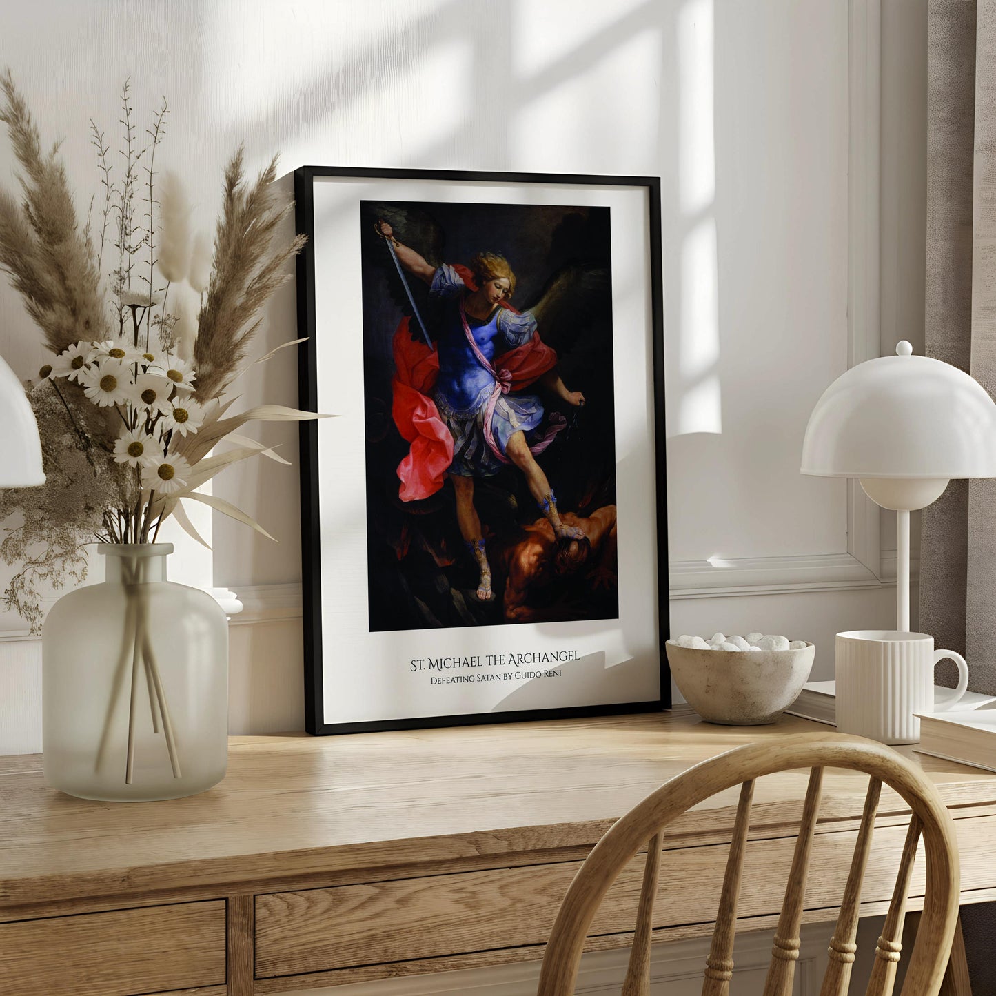 a framed painting of St. Michael the Archangel, a religious figure, standing in a dramatic pose with a sword in his hand. The painting is displayed on a white wall above a desk with various items on it, including a notebook, a cup, and a vase.