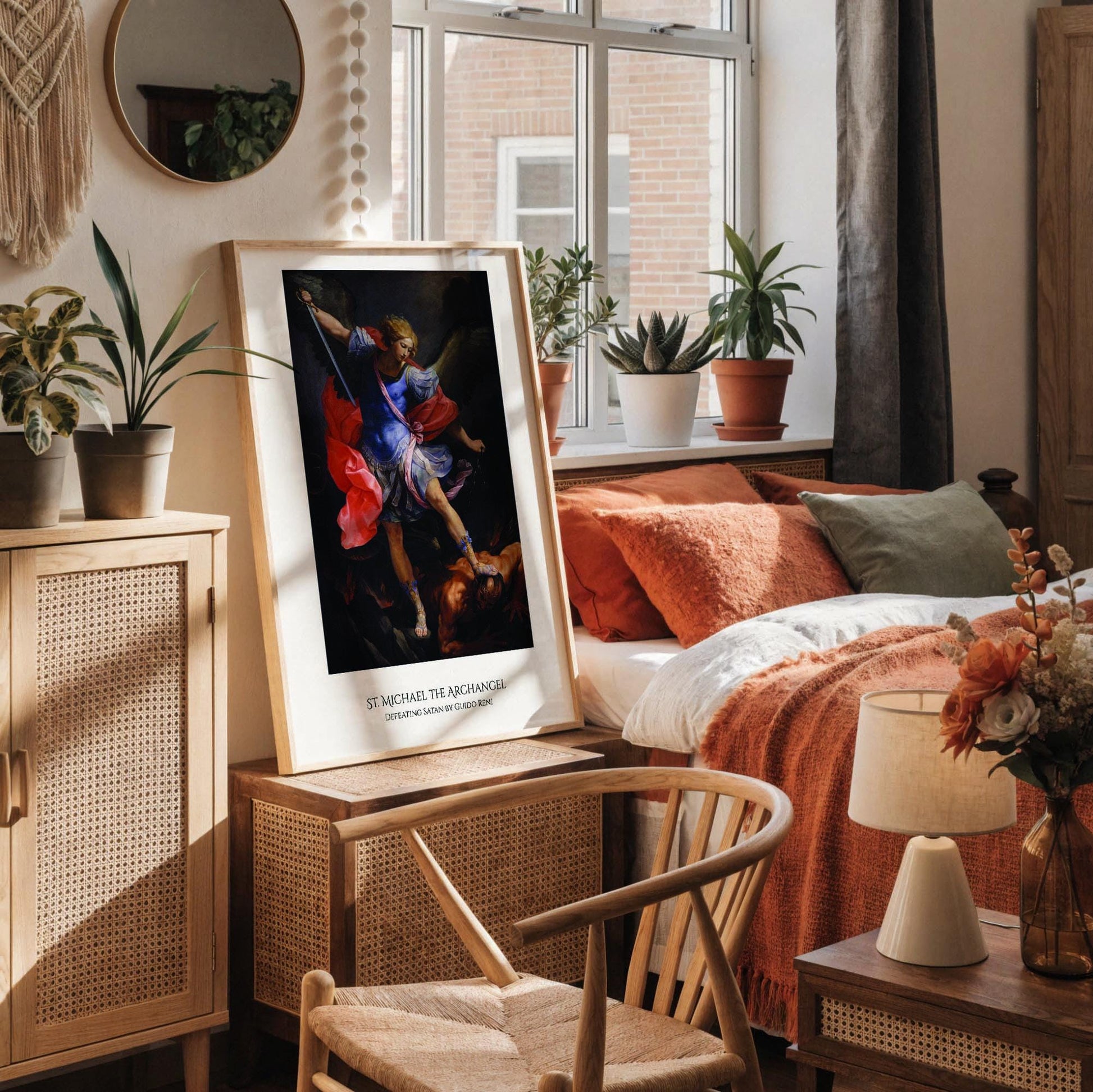 a framed painting of St. Michael, the archangel, with a white desk in the foreground. The painting depicts St. Michael holding a sword and a shield, with a figure in the background. The desk has various items on it, including a cup, a notebook, and a vase with dried flowers.