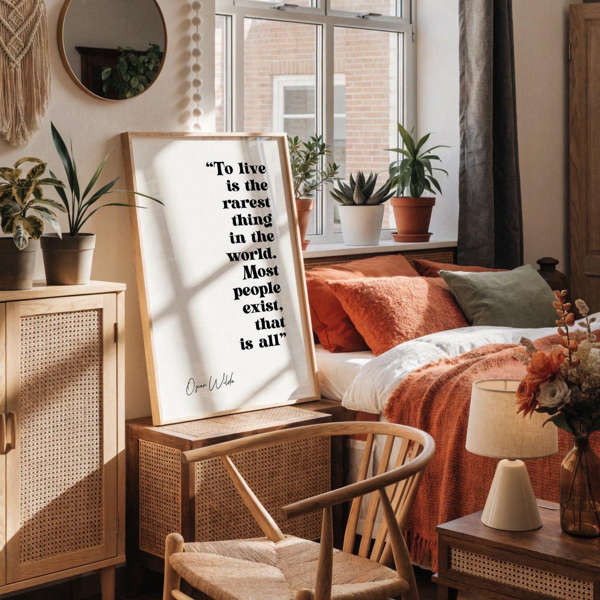 a framed quote by Oscar Wilde, "To live is the rarest thing in the world. Most people exist, that is all", displayed on a white wall above a desk with various items on it, including a vase with dried flowers, a notebook, a cup, and a pen holder.