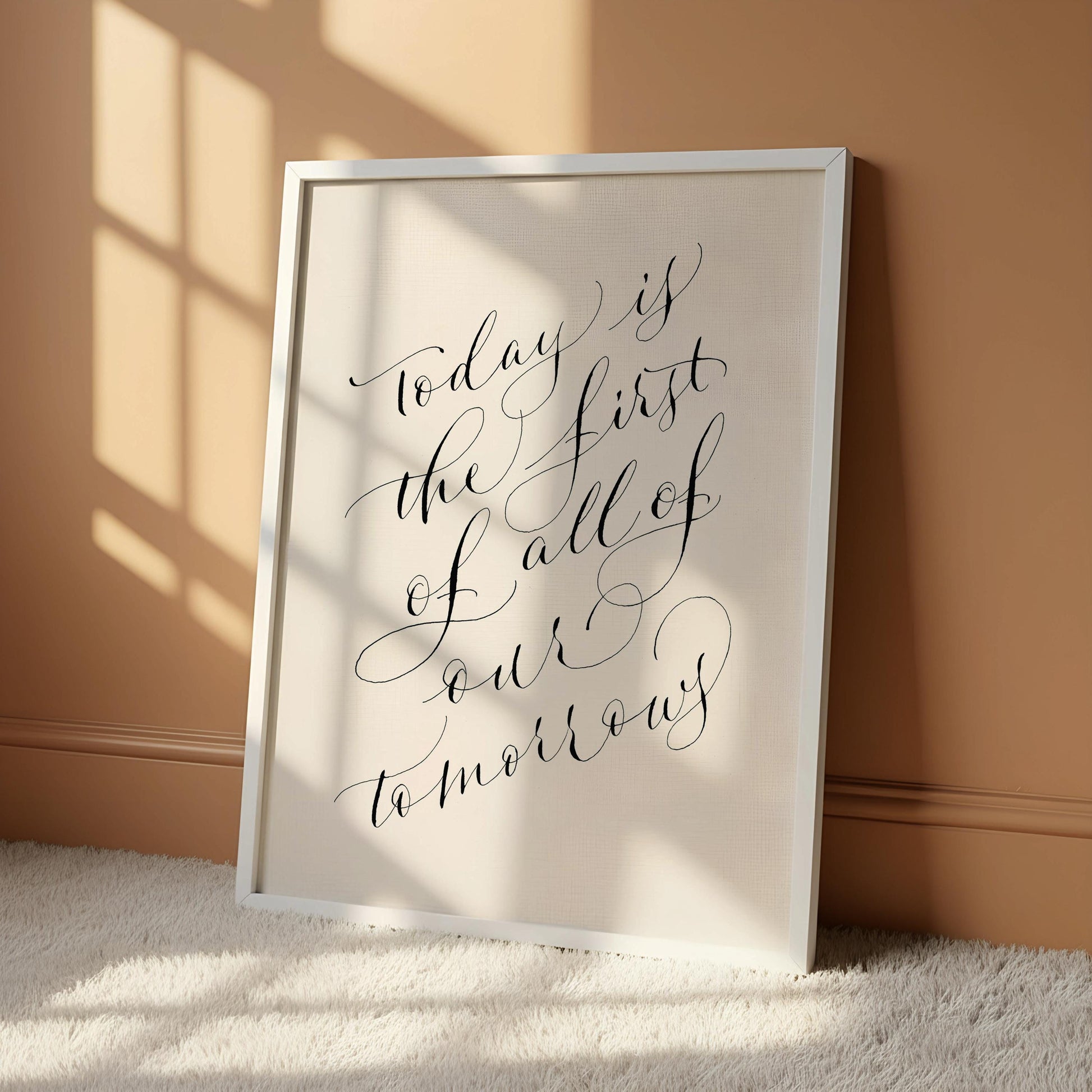 a framed quote with a black frame, placed on a white desk with various decorative items such as a vase with dried flowers, a small white candle, and a white mug.
