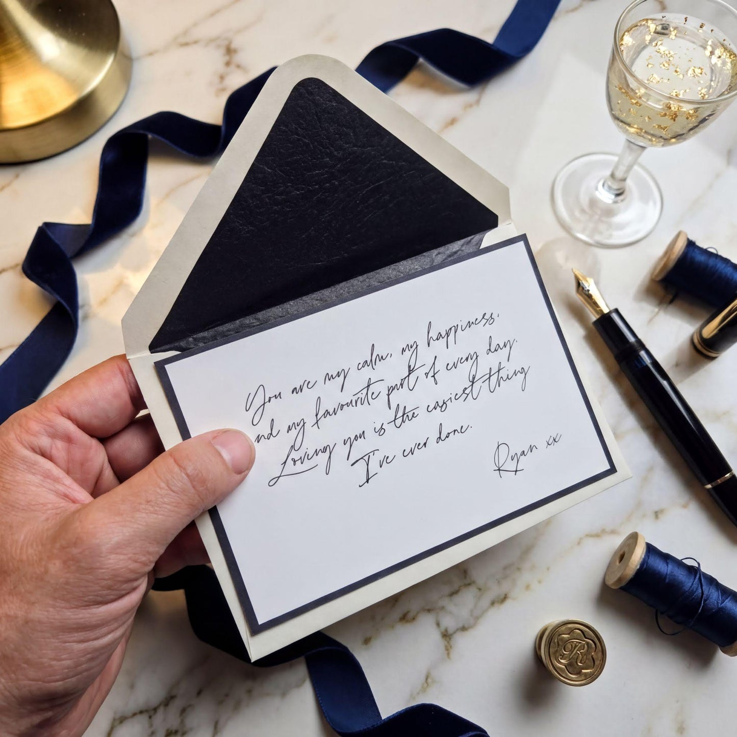A hand holding an open card with a handwritten message, accompanied by a glass of wine, a pen, and some blue ribbons on a marble surface.