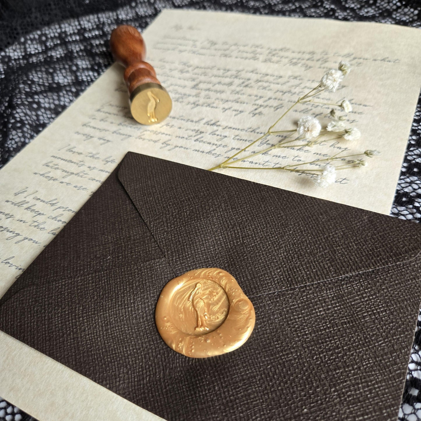 a close-up view of a black envelope with a gold wax seal, a handwritten letter, and a small bouquet of white flowers.