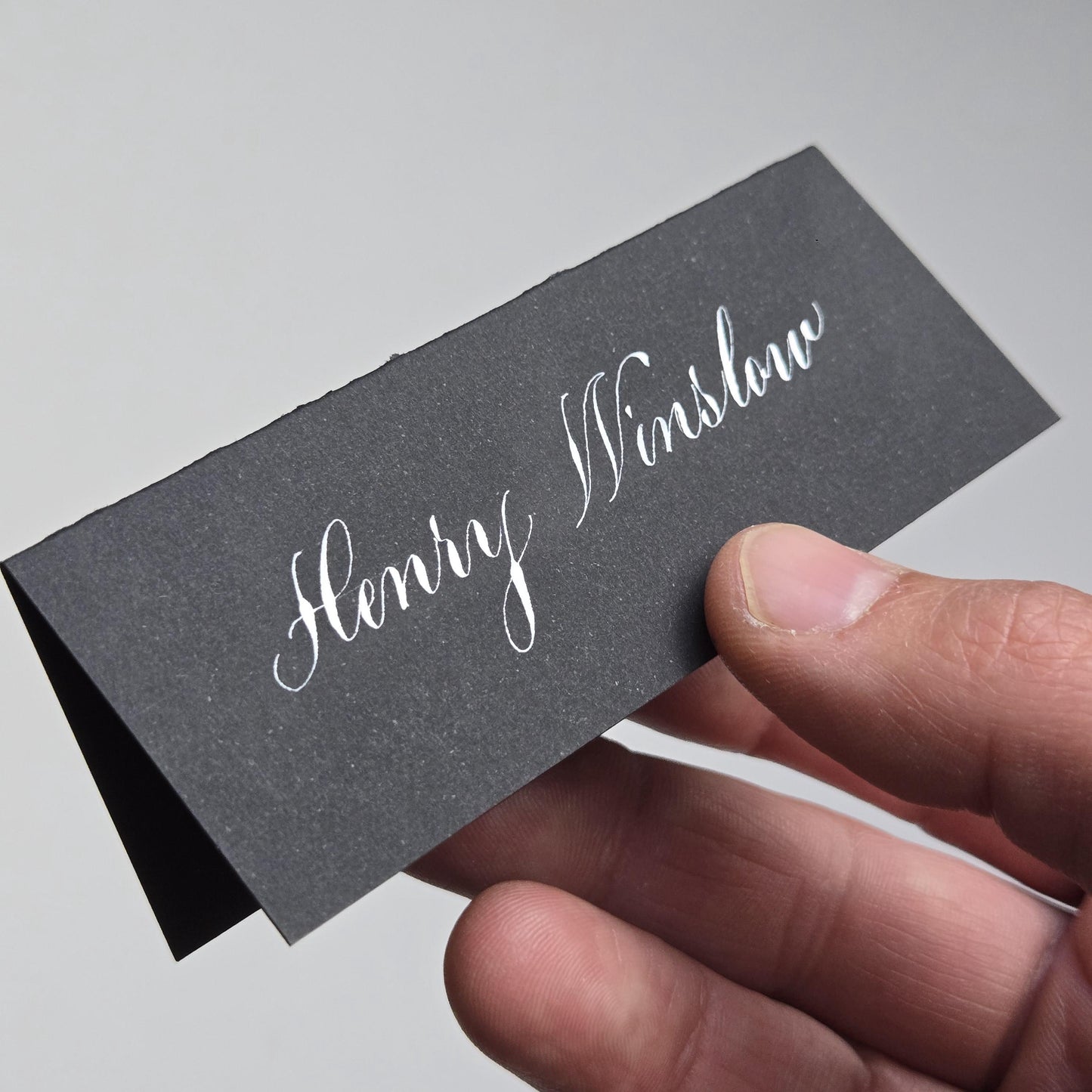 A hand holding a black rectangular card with the name "Henry Winslow" written on it in elegant cursive script.