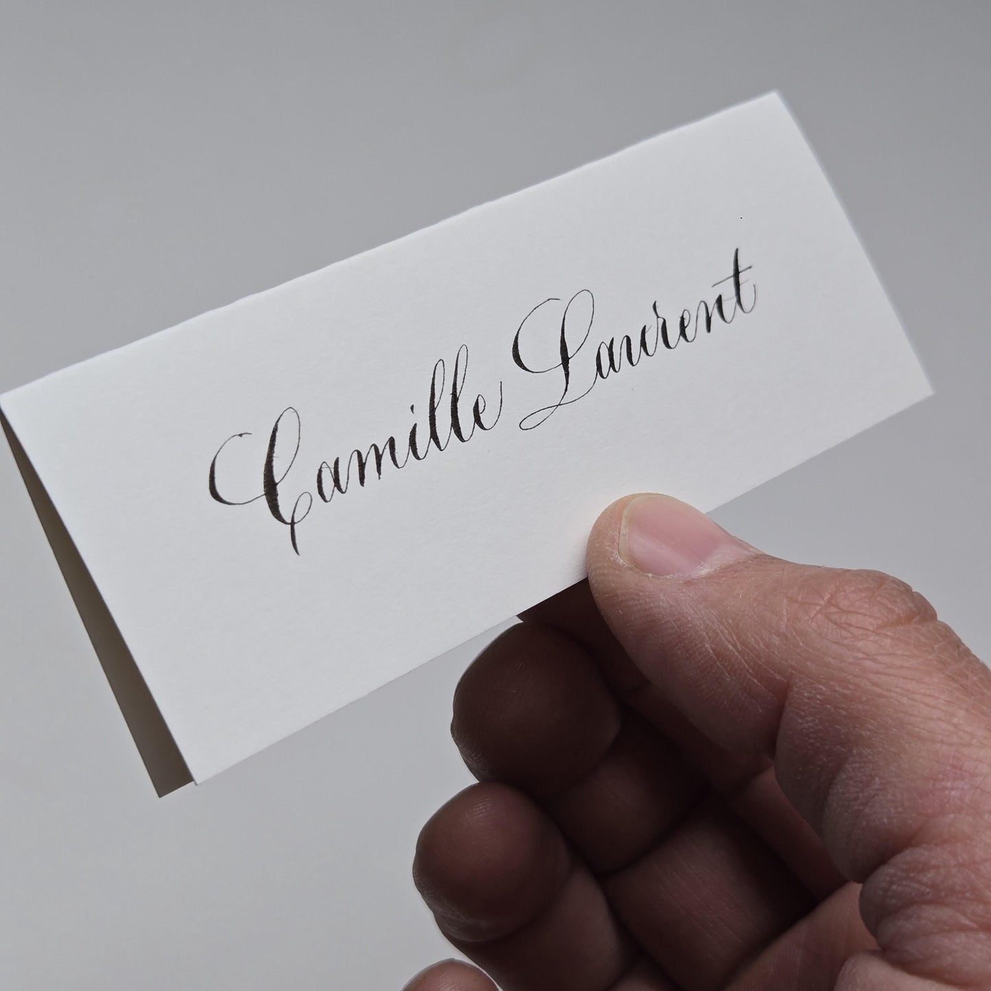 A hand holding a white card with the name "Camille Laurent" written on it.