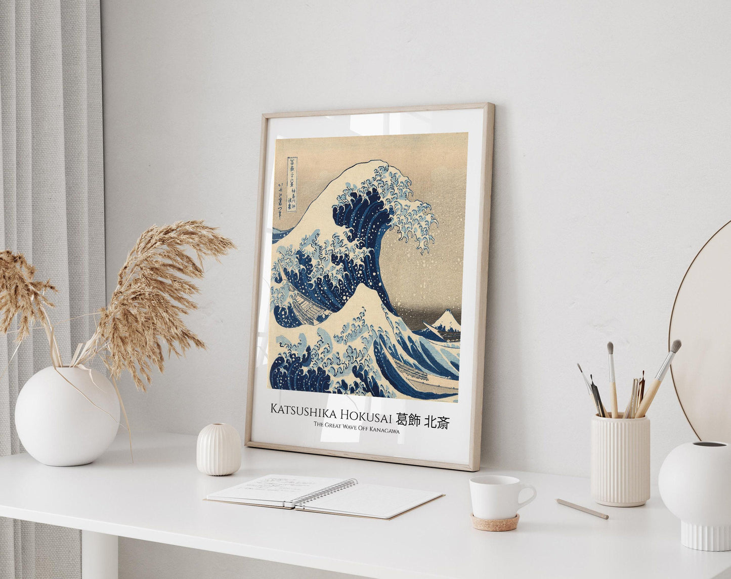 a framed poster of the famous Japanese woodblock print "The Great Wave off Kanagawa" by the artist Katsushika Hokusai, displayed on a white wall above a desk with various items on it.