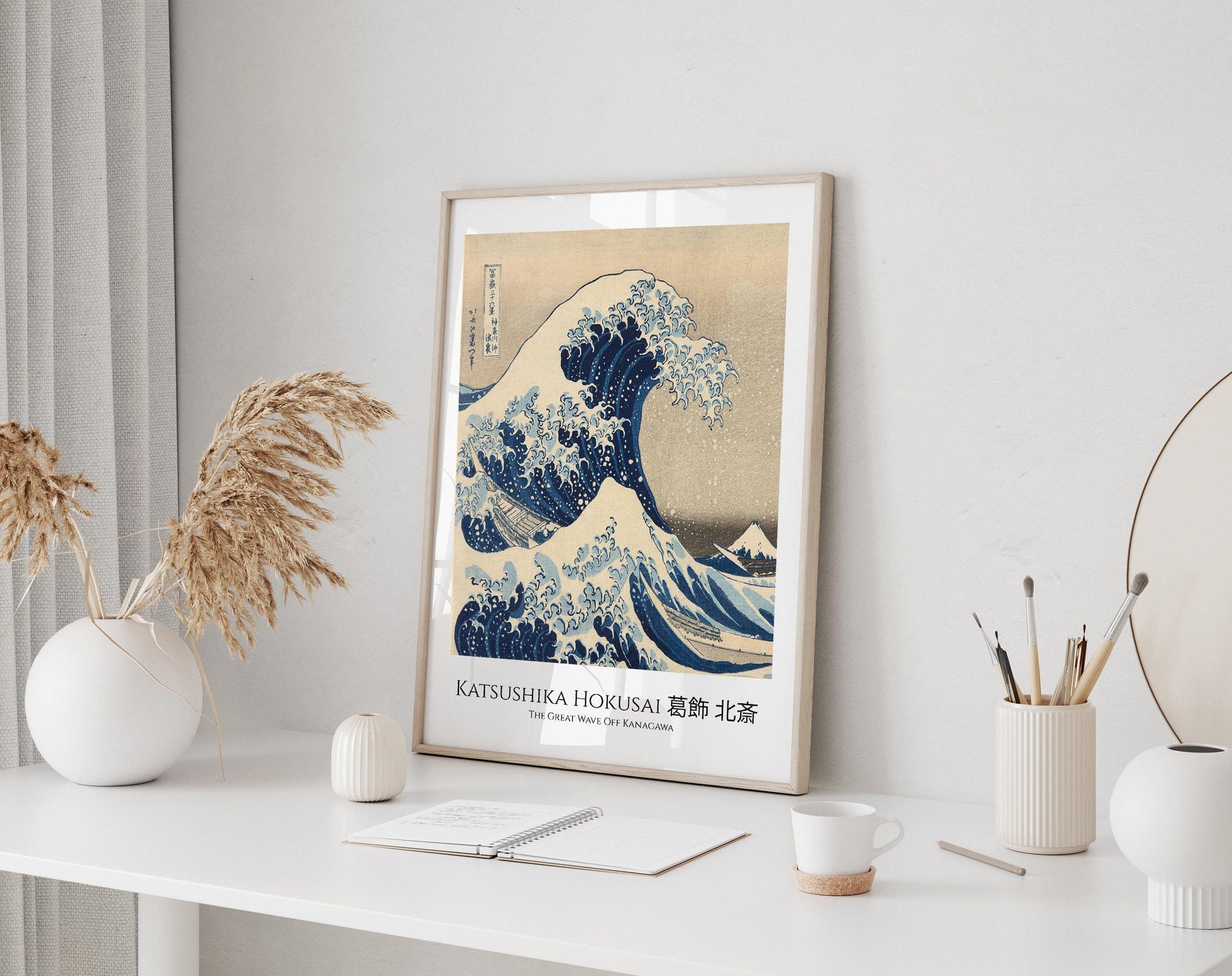 a framed poster of the famous Japanese woodblock print "The Great Wave off Kanagawa" by the artist Katsushika Hokusai, displayed on a white wall above a desk with various items on it.