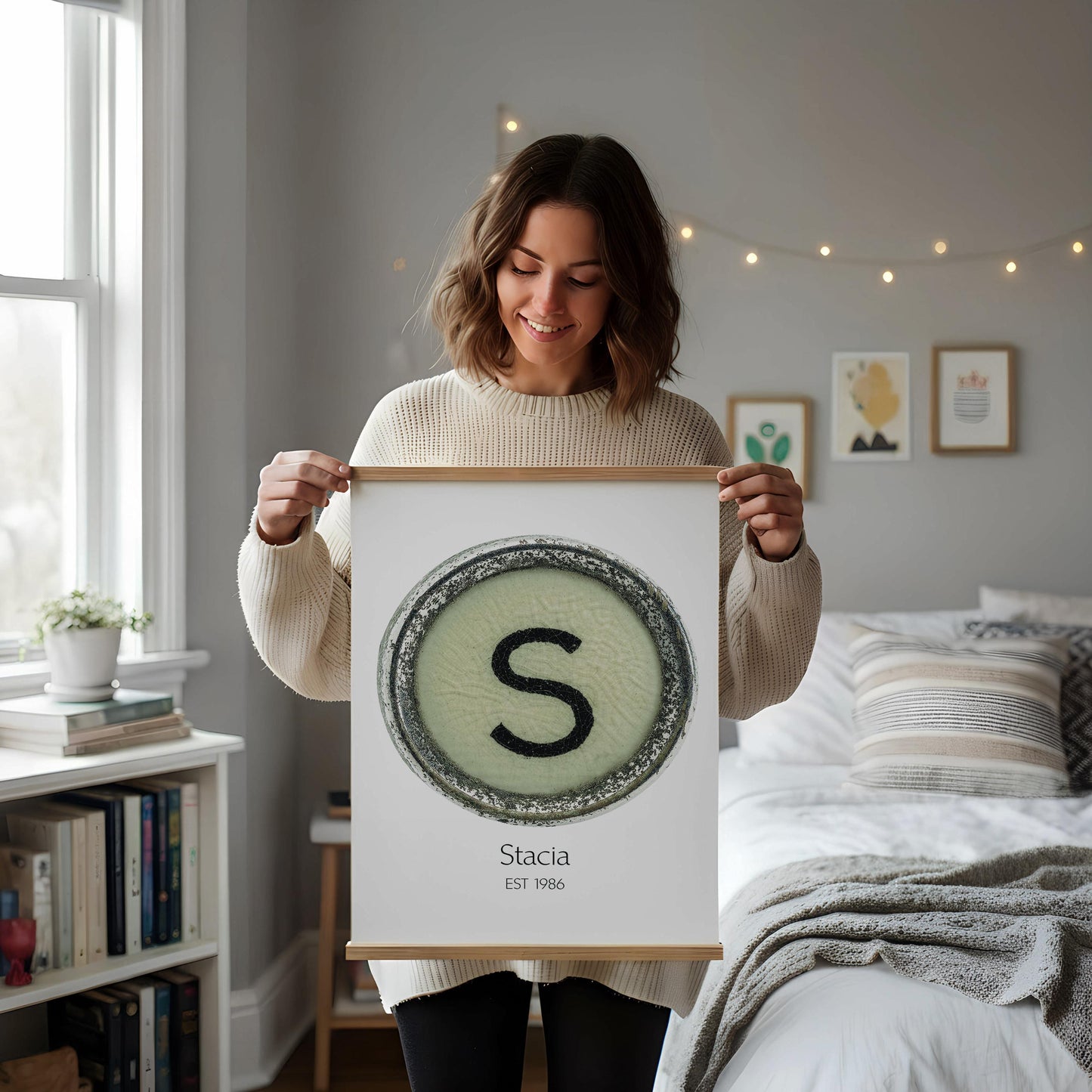 a framed artwork with a large letter 'S' on a white background, placed on a white desk. The desk also has a notebook, a cup, and a vase with dried flowers.
